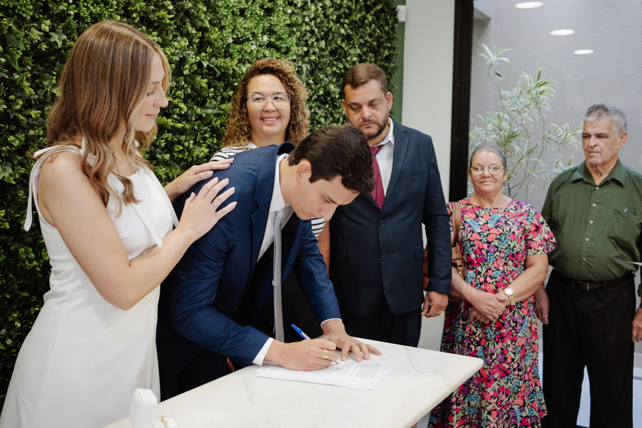 Noivo assina documentos do casamento civil no Cartório de São José dos Campos sob o olhar carinhoso da noiva grávida e familiares. Registro profissional de fotografia de casamento civil em SJC com foco na emoção e nos detalhes da cerimônia.