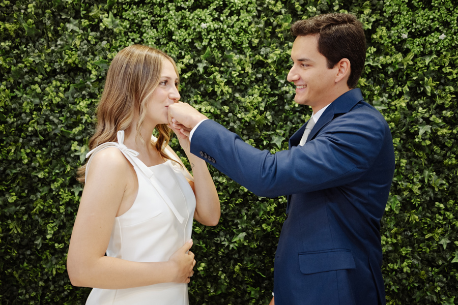 Noivo beija a mão da noiva grávida em frente a um painel de folhagens após cerimônia de casamento civil em São José dos Campos. Fotografia profissional em SJC destacando o carinho e a emoção de Ana Luiza e Felipe Antônio na espera pela pequena Laura.