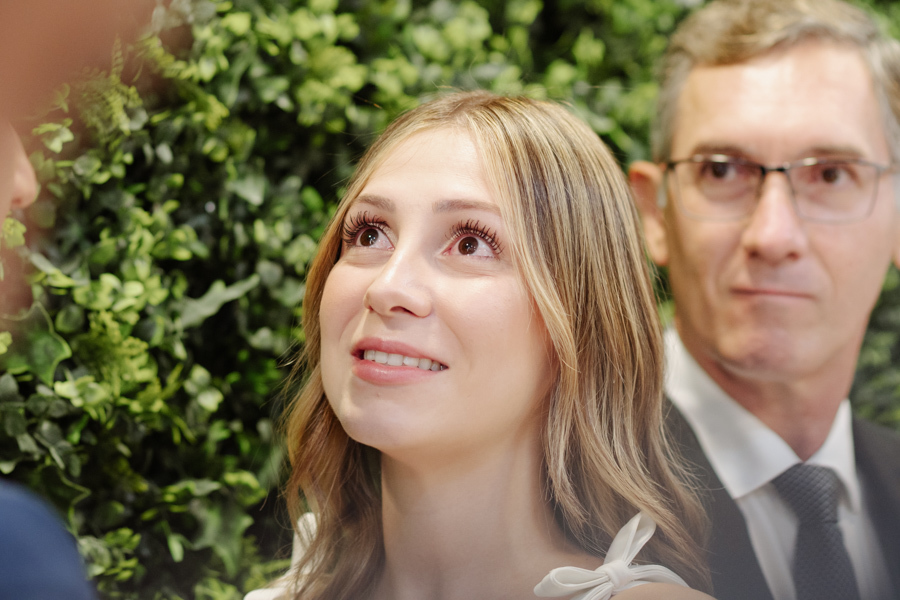Retrato em close da noiva Ana Luiza com olhar emocionado e sorridente durante cerimônia no Cartório de São José dos Campos. Fotografia de casamento civil em SJC que captura a sensibilidade e a felicidade da noiva em frente ao painel de folhagens.