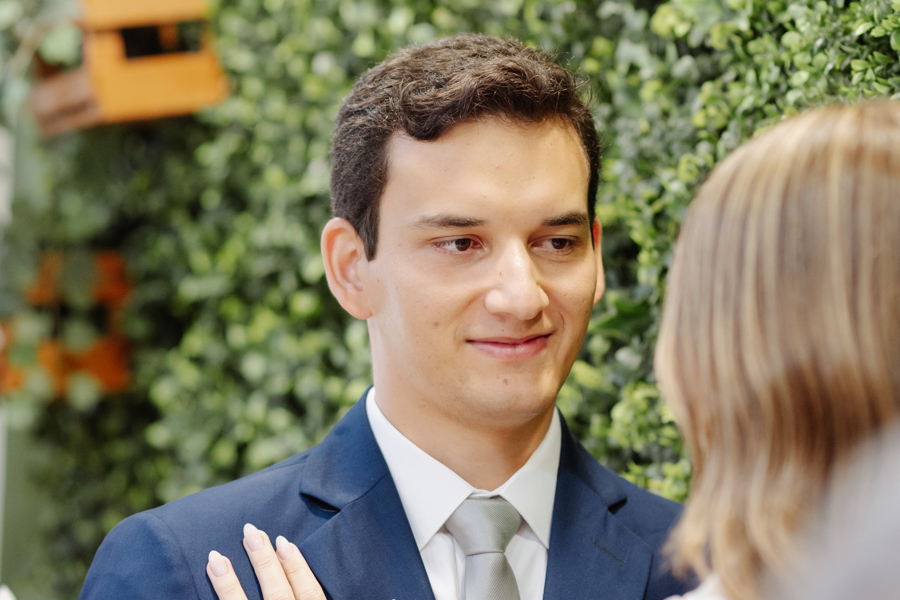 Retrato em close do noivo Felipe Antônio com expressão de felicidade e olhar sereno durante cerimônia no Cartório de São José dos Campos. Fotografia de casamento civil em SJC que destaca a emoção do noivo em frente ao painel de folhagens.