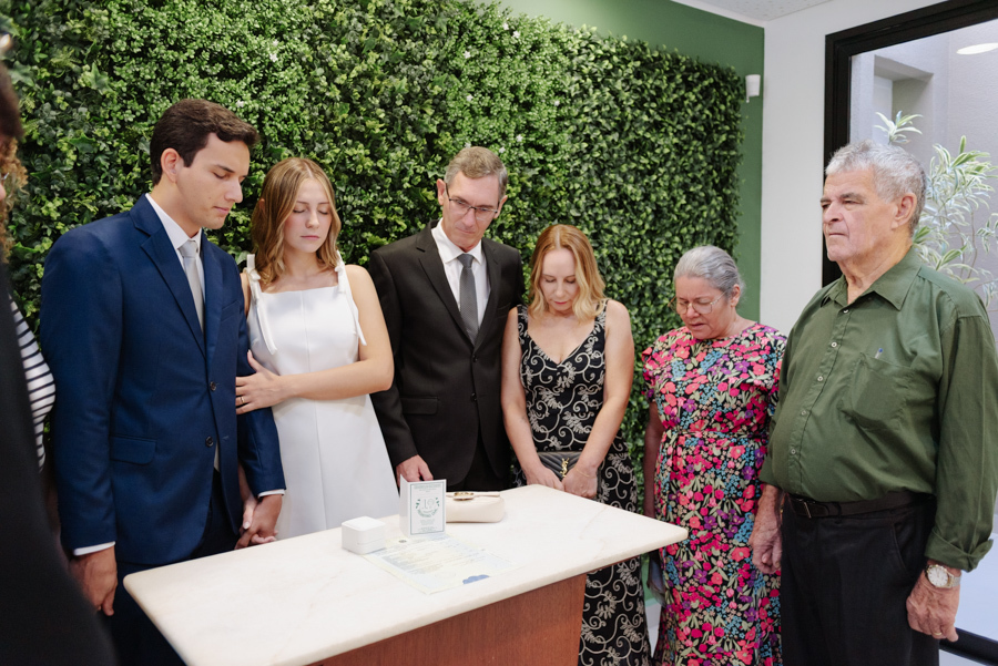 Casal de noivos e familiares em momento de oração ou silêncio com cabeças baixas durante cerimônia no Cartório de São José dos Campos. Fotografia de casamento civil em SJC que registra a espiritualidade e o respeito da família de Ana Luiza e Felipe Antôni