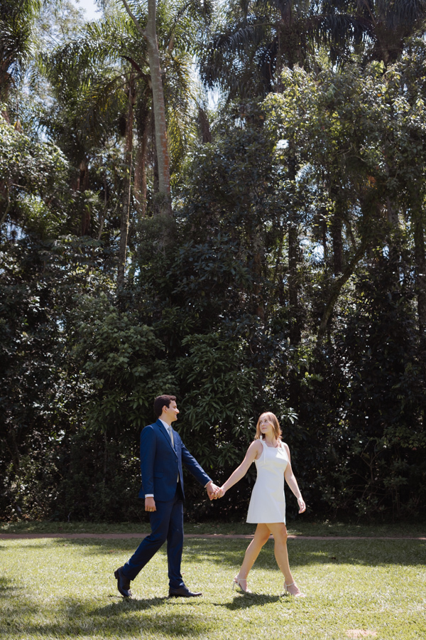Casal caminha de mãos dadas pelo gramado ensolarado do Parque da Cidade em São José dos Campos. Fotografia de ensaio de casamento civil em SJC destacando Ana Luiza e Felipe Antônio em meio à natureza exuberante do Parque Roberto Burle Marx.