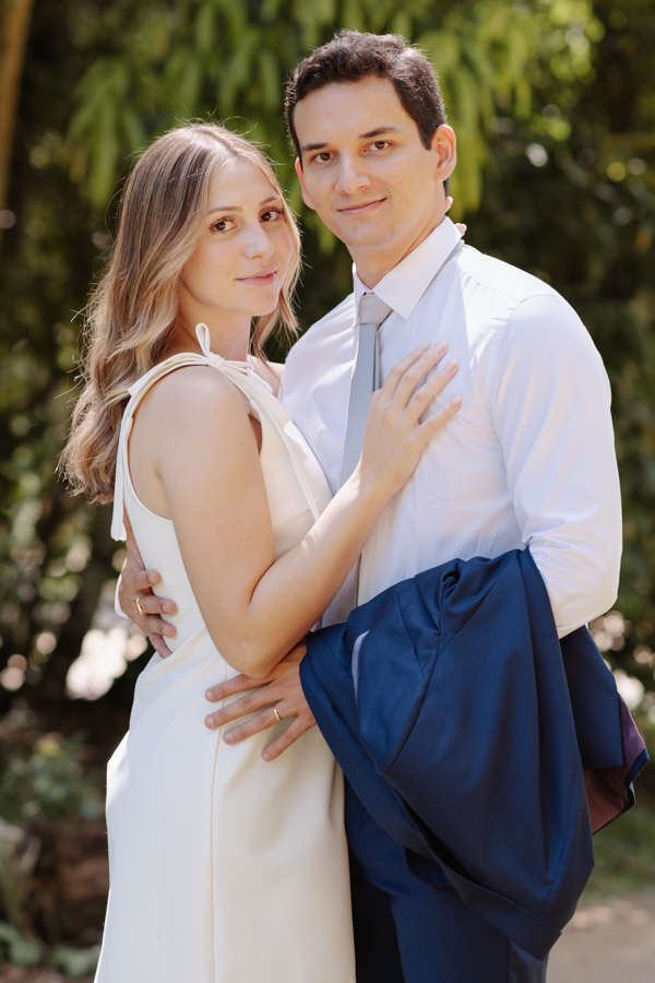 Retrato frontal dos noivos Ana Luiza e Felipe Antônio em ensaio externo. A noiva, grávida, usa vestido branco e o noivo segura seu paletó azul. Fotografia de casamento civil em SJC capturando a serenidade do casal em meio à natureza de São José dos Campos
