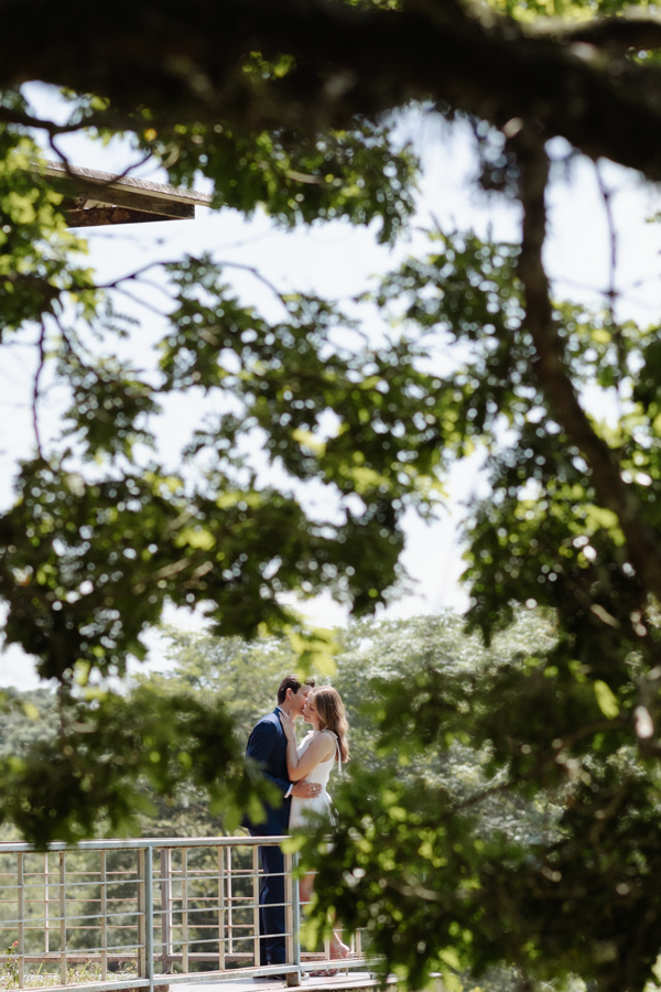 Casal de noivos se abraça e troca carinhos em deck de madeira, emoldurados pelas folhas das árvores. Fotografia de ensaio de casamento civil em SJC capturando o romance de Ana Luiza e Felipe Antônio em meio à natureza de São José dos Campos.