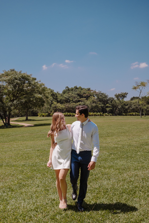 Ana Luiza e Felipe Antônio caminham sorridentes pelo gramado ensolarado do Parque da Cidade em São José dos Campos. Fotografia de ensaio de casamento civil em SJC que captura a felicidade do casal em meio à natureza do Parque Roberto Burle Marx.