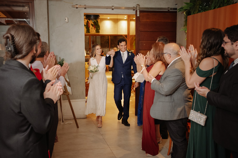 Entrada triunfal dos noivos Ana Luiza e Felipe Antônio no restaurante Auá Gastronomia em SJC. Fotografia de recepção de casamento civil em São José dos Campos com convidados aplaudindo o casal em um momento de celebração e muita alegria.