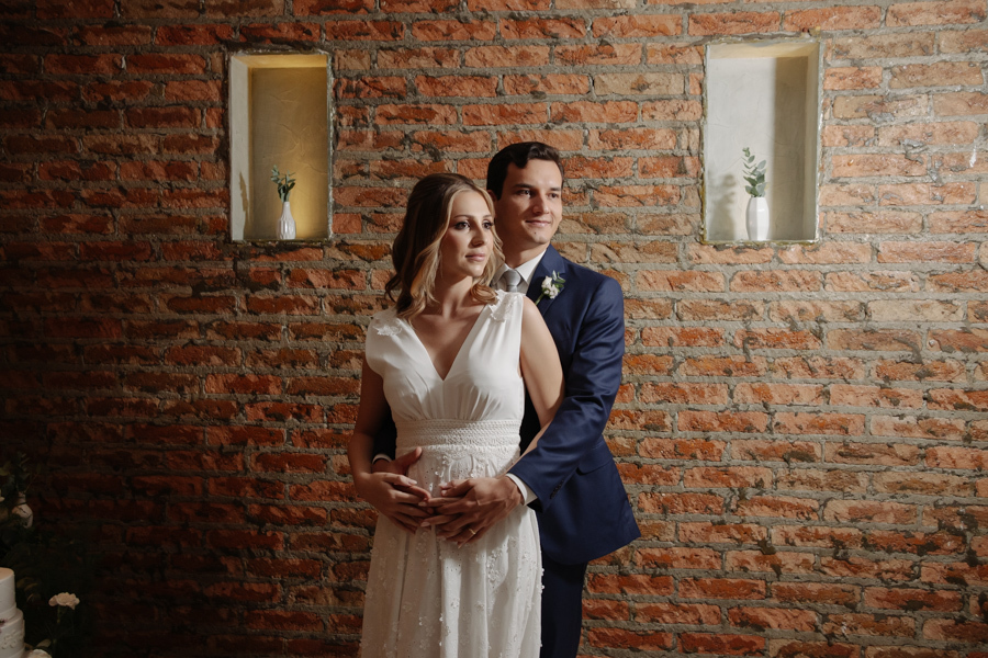 Retrato dos noivos Ana Luiza e Felipe Antônio abraçados em frente a uma parede de tijolos rústicos no restaurante Auá Gastronomia. Fotografia de recepção de casamento civil em SJC, destacando a elegância do casal e a doce espera pela pequena Laura.