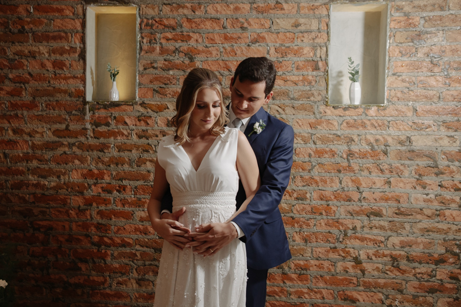 Felipe Antônio abraça Ana Luiza por trás enquanto olham para a barriga de grávida da noiva no restaurante Auá Gastronomia. Fotografia de casamento civil em SJC que destaca a doçura e a cumplicidade do casal à espera da pequena Laura.