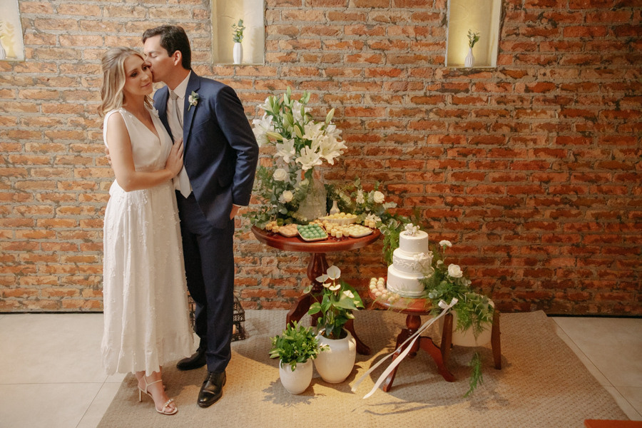 Felipe Antônio beija o rosto de Ana Luiza ao lado da mesa de doces e do bolo de casamento no restaurante Auá Gastronomia. Fotografia de recepção de casamento civil em SJC que destaca o carinho do casal e a decoração elegante em São José dos Campos.