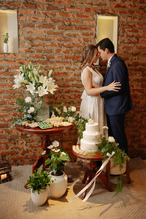 Noivos encostam as testas em momento de carinho ao lado da mesa de doces e do bolo de casamento no restaurante Auá Gastronomia. Fotografia de recepção de casamento civil em SJC que destaca a sintonia de Ana Luiza e Felipe Antônio em São José dos Campos.
