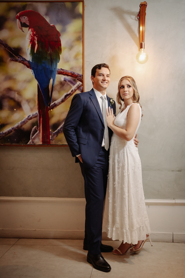Noivos Ana Luiza e Felipe Antônio posam abraçados em frente a um quadro de arara no restaurante Auá Gastronomia. Fotografia de recepção de casamento civil em SJC que destaca a elegância e a felicidade do casal em São José dos Campos.