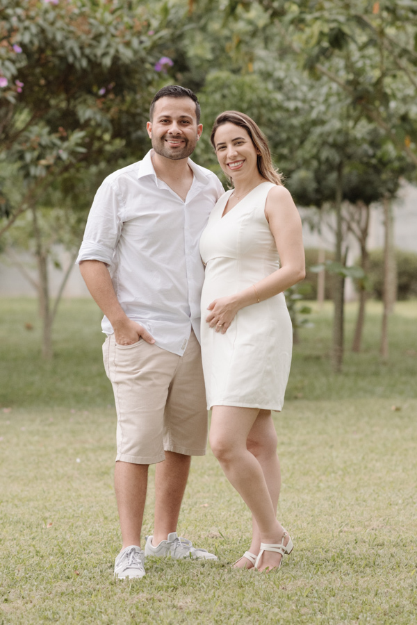 Uma foto de um casal sorridente em um parque. O homem veste camisa branca e bermuda bege; a mulher, grávida, usa um vestido branco curto e acaricia a barriga. Eles estão em pé sobre o gramado, cercados por árvores ao fundo sob luz natural suave.