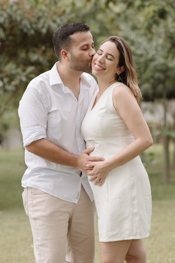Casal sorridente em ensaio gestante ao ar livre. O homem, de camisa branca e bermuda bege, beija o rosto da mulher, que usa vestido branco e segura a barriga. Fundo com árvores e gramado sob luz natural suave.