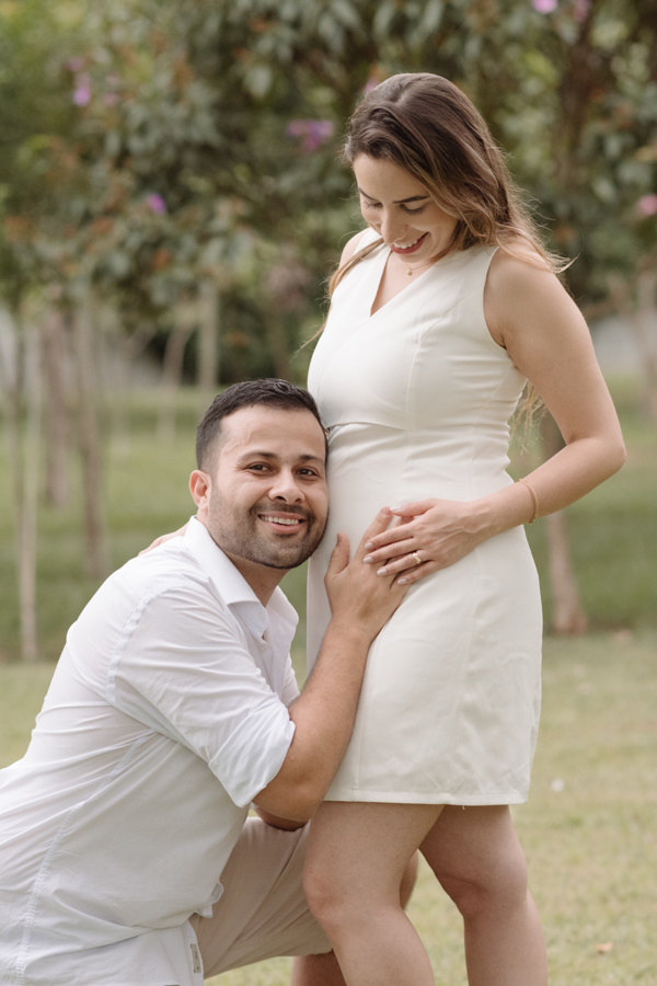 Homem sorridente ajoelhado encosta o ouvido na barriga da mulher grávida em um parque. Ambos vestem branco. A gestante olha para baixo com carinho enquanto ele segura sua barriga. Fundo arborizado com luz natural suave.