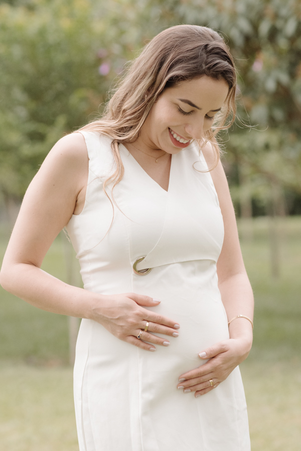 Gestante sorridente em plano médio, usando vestido branco, olha para baixo com carinho enquanto segura suavemente a barriga com as duas mãos. Ela está em um jardim com fundo desfocado e iluminação natural suave.