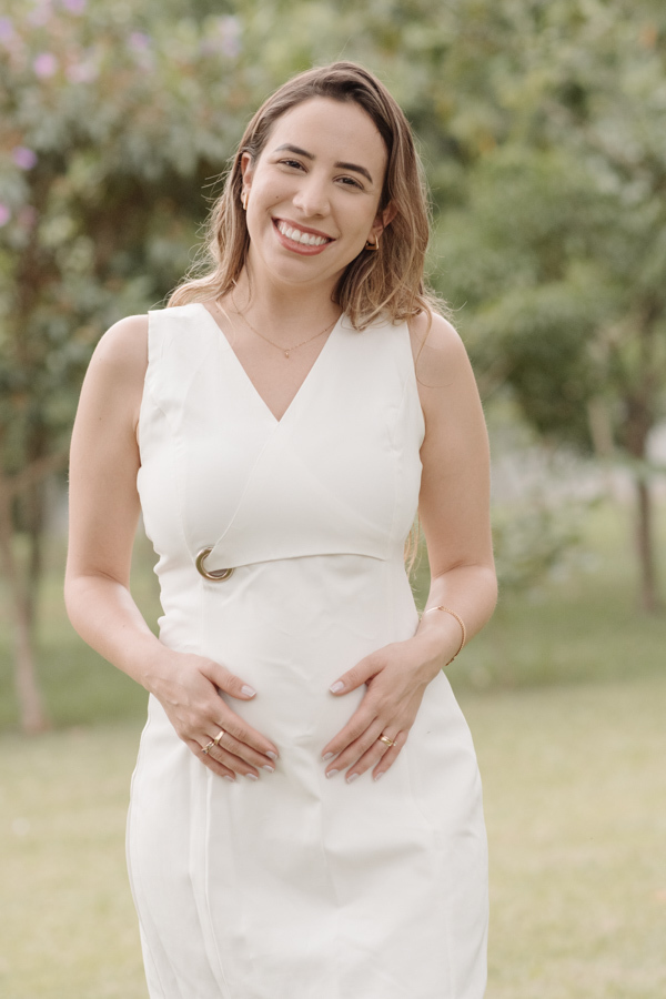 Gestante sorridente em plano médio, usando vestido branco curto, posa de frente em um jardim. Ela segura a barriga com as duas mãos em um gesto de carinho. O fundo arborizado está suavemente desfocado, destacando a felicidade da mãe sob luz natural.