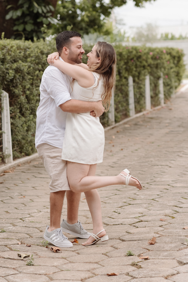 Casal se abraça sorrindo em uma rua de paralelepípedos. O homem levanta a mulher, que dobra uma das pernas para trás. Ambos vestem branco e bege. Ao fundo, uma cerca viva verde e árvores sob luz natural suave eternizam o momento de celebração.