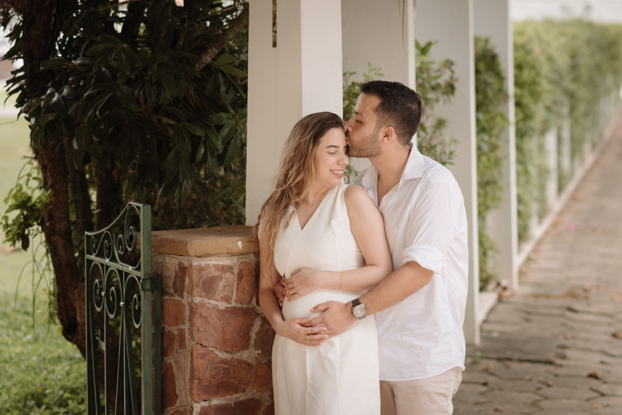 Homem beija a testa da gestante em momento afetuoso ao ar livre. Ambos vestem branco e estão abraçados, com as mãos sobre a barriga dela. O cenário inclui colunas brancas e vegetação, capturando a serenidade e a conexão do casal sob luz natural.