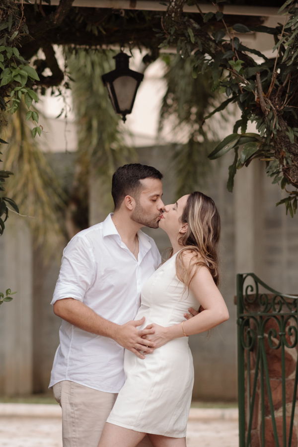 Casal se beija apaixonadamente sob um arco de folhagens naturais. Ambos vestem roupas claras e estão com as mãos na barriga da gestante. O cenário inclui uma luminária rústica e portão de ferro, criando uma atmosfera romântica e acolhedora.