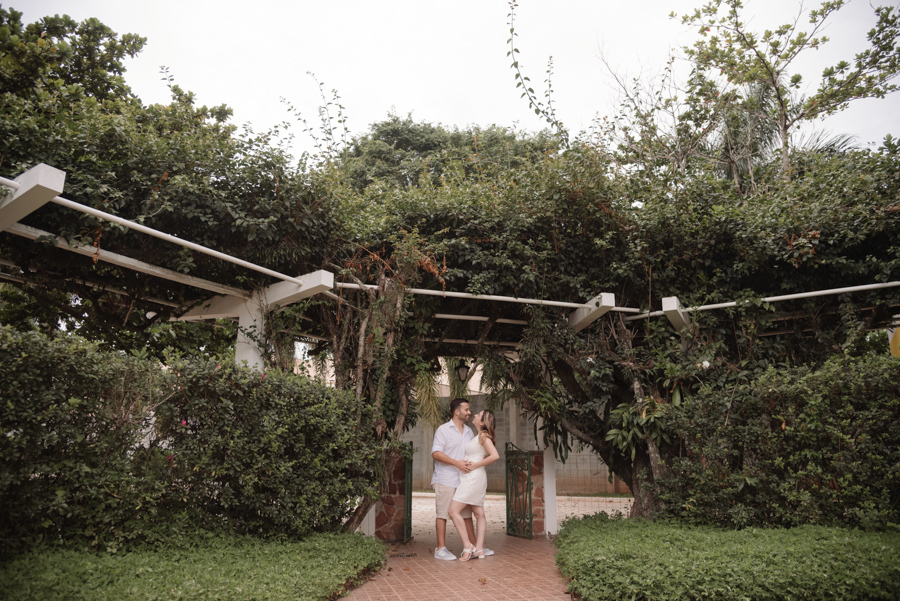 Casal posa sorridente sob um grande pergolado coberto por densa vegetação verde. Ambos vestem roupas claras e estão abraçados em um caminho de pedras cercado por arbustos, em um registro aberto que destaca a harmonia entre o casal e a natureza.