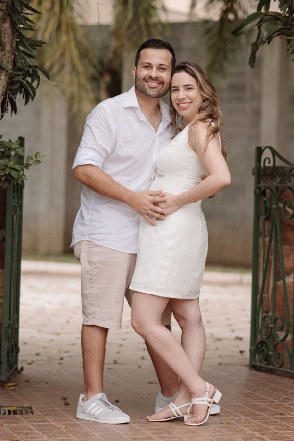 Casal sorridente posa abraçado em frente a um portão de ferro rústico. O homem, de camisa branca, envolve a barriga da gestante, que usa vestido branco. O cenário é arborizado, com luz natural suave realçando a alegria e união da nova família.