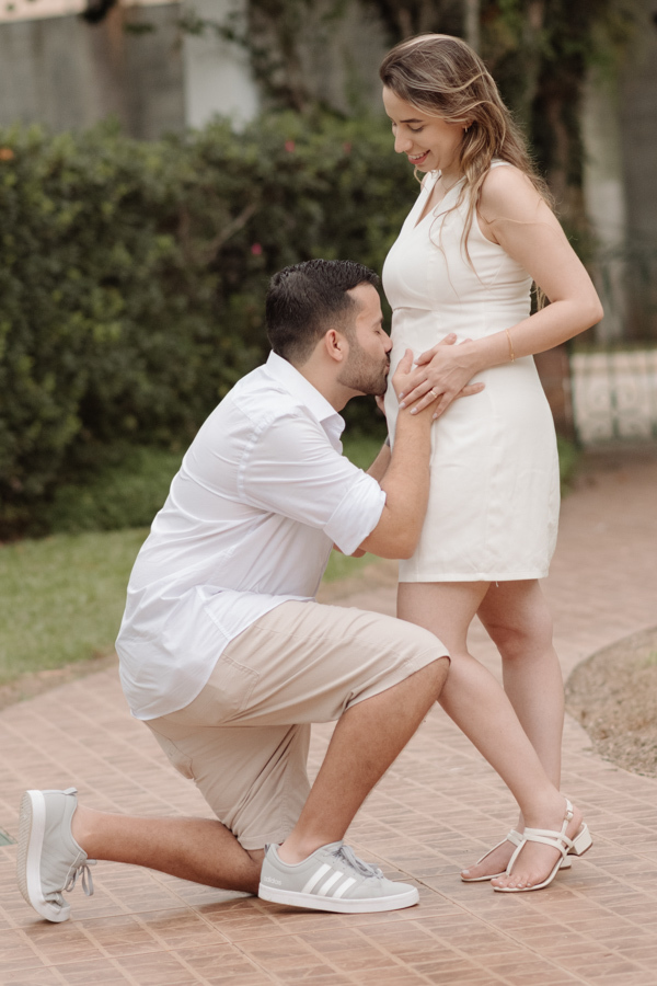 Homem ajoelhado beija carinhosamente a barriga da gestante em um caminho de pedras. Ambos vestem roupas claras e sorriem. O fundo apresenta vegetação verde e luz natural, destacando a ternura e a conexão do casal à espera do bebê.