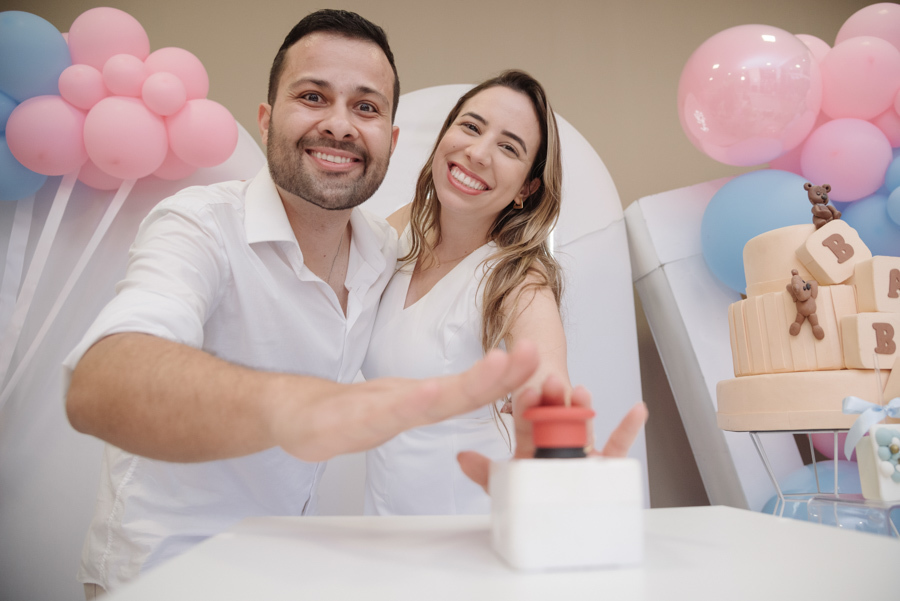 Casal sorridente em close-up, prestes a apertar o botão de ativação da grande revelação. Diego e Tamires celebram esse momento de muita expectativa em um cenário alegre, decorado com balões azuis e rosa, ansiosos para descobrir o destino dessa nova famíli