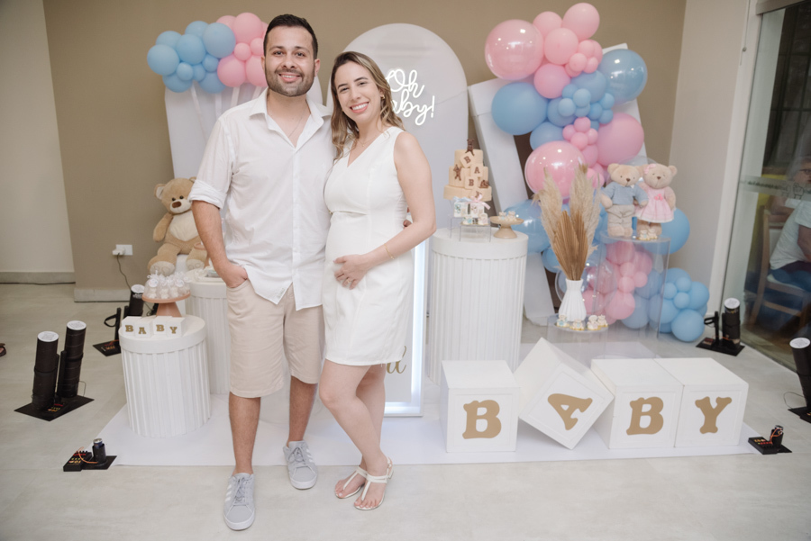 Casal sorridente posa à frente da decoração de chá revelação, com balões azuis e rosa, ursinhos e letreiro luminoso. Diego e Tamires celebram esse capítulo especial, com Tamires segurando a barriga em um momento de pura expectativa e alegria.