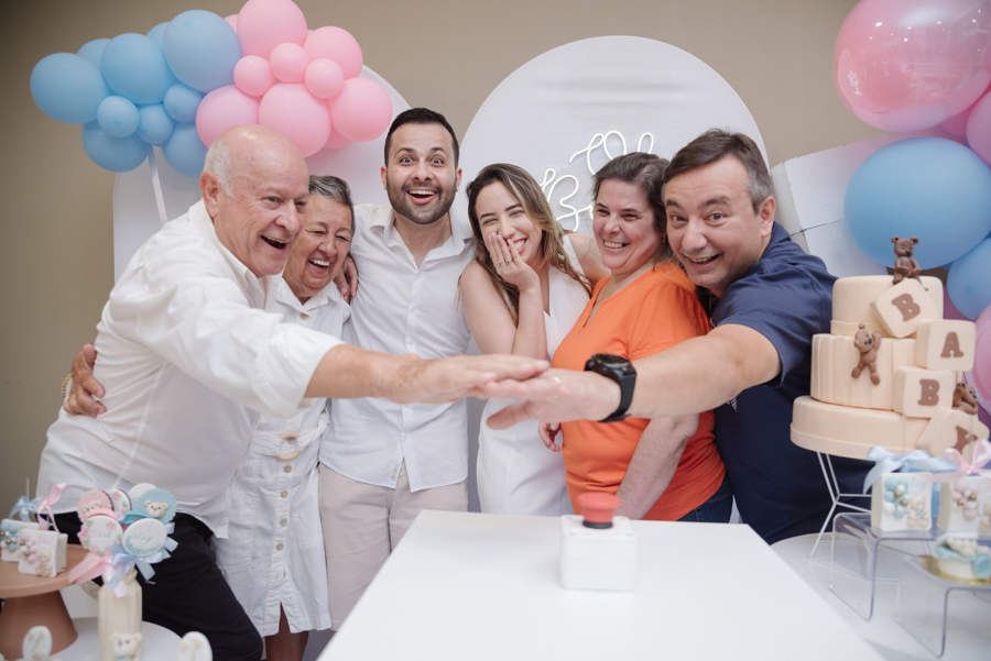 Grupo de familiares e o casal sorridente reunidos em volta de um botão de ativação, em um momento de grande expectativa. O cenário decorado com balões azuis e rosa e um bolo temático prepara a grande revelação sob um clima de pura alegria e união.