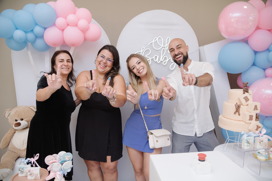 Quatro convidados sorridentes mostram os dedos sujos de tinta rosa e azul em frente à decoração do chá revelação. O momento de descontração e expectativa de Diego e Tamires é registrado entre balões, ursinhos e o botão que revelará o destino da família.