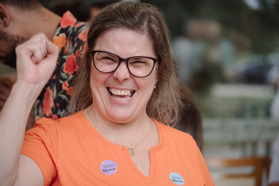 Convidada sorridente vibra de alegria durante o chá revelação de Diego e Tamires. Ela usa blusa laranja com adesivos 