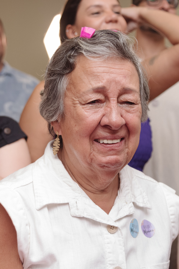 Uma convidada idosa se emociona no chá revelação de Diego e Tamires, com lágrimas nos olhos e um sorriso sincero. Ela usa adesivos de aposta e tem um papel picado rosa no cabelo, capturando a pura emoção desse capítulo inesquecível da nova família.