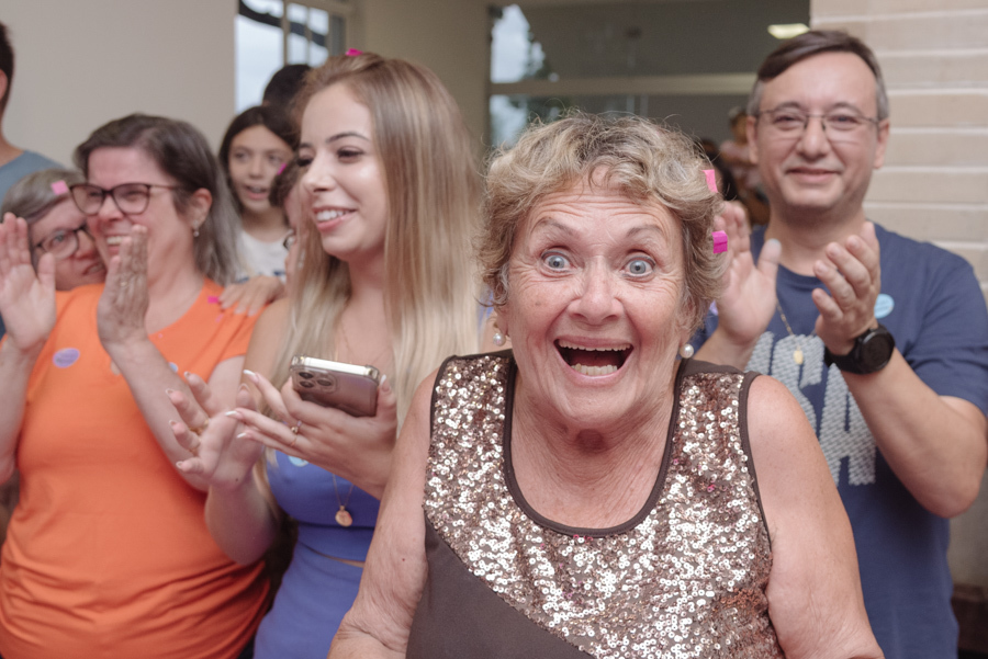 Uma convidada vibra com uma expressão de choque e pura alegria durante a grande revelação de Diego e Tamires. Ao fundo, amigos e familiares aplaudem o momento mágico em que o destino da nova família se tingiu de rosa sob muita expectativa e emoção.