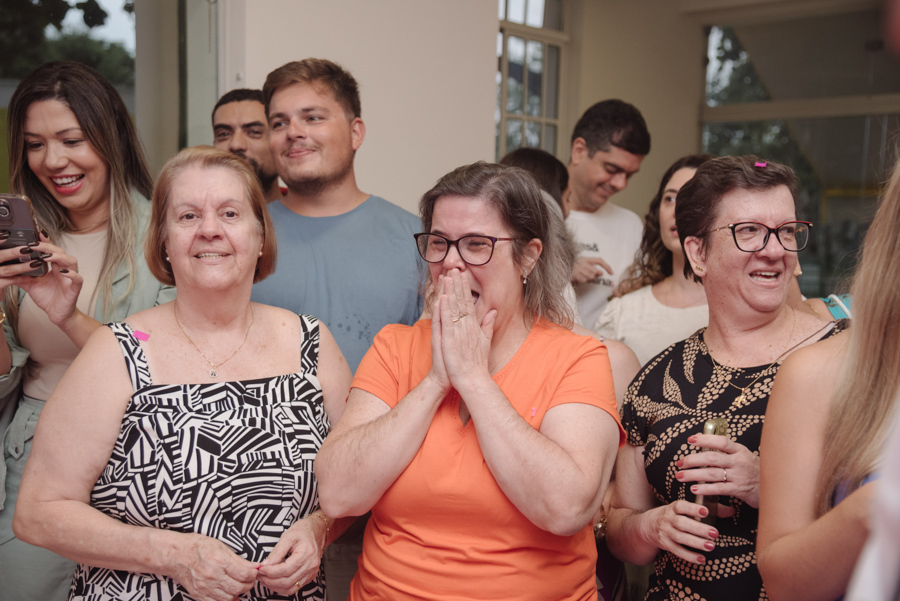 Convidadas emocionadas reagem com alegria e surpresa à revelação do sexo do bebê de Diego e Tamires. No centro, uma mulher de laranja cobre o rosto, capturando o momento em que a vida da nova família se tingiu de rosa sob muita expectativa e felicidade.