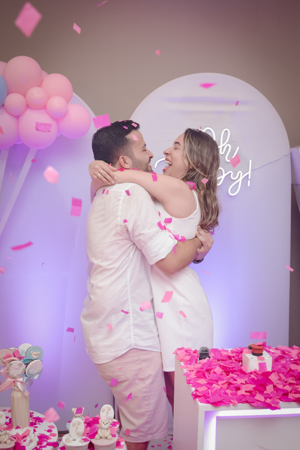 Diego e Tamires se abraçam rindo em meio à chuva de papéis rosa. O momento mágico revela que a vida da nova família se tingiu de rosa, capturando a felicidade contagiante do casal em um cenário decorado com balões e letreiro luminoso 