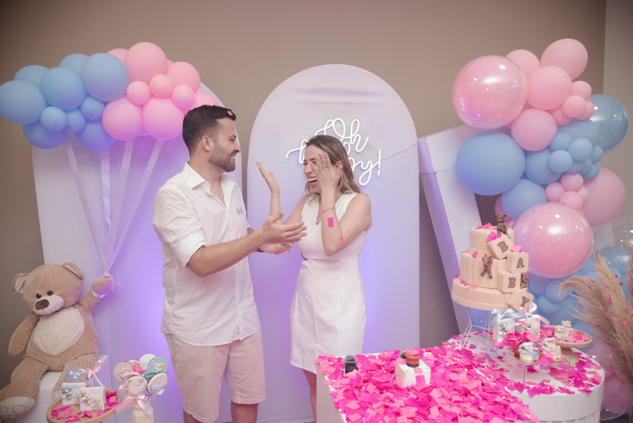 Diego e Tamires rindo e celebrando após a grande revelação, com papéis rosa espalhados pelo cenário. O casal compartilha a alegria desse momento mágico em que a vida da nova família se tingiu de rosa, cercados por uma decoração encantadora e festiva.