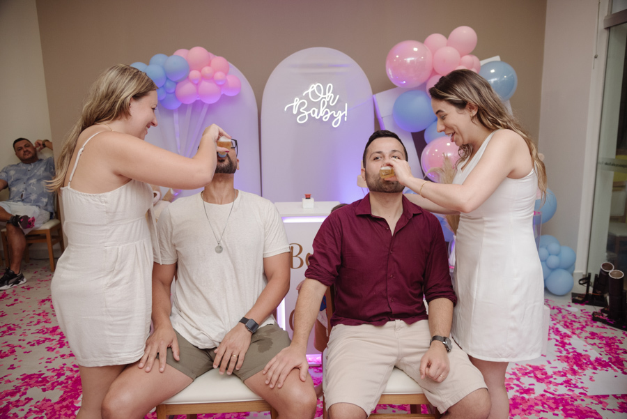 Tamires ajuda um convidado a beber um shot, enquanto Diego também participa da dinâmica após a revelação. O chão está coberto de papéis rosa, celebrando com descontração o destino da família em um cenário festivo com balões e letreiro 
