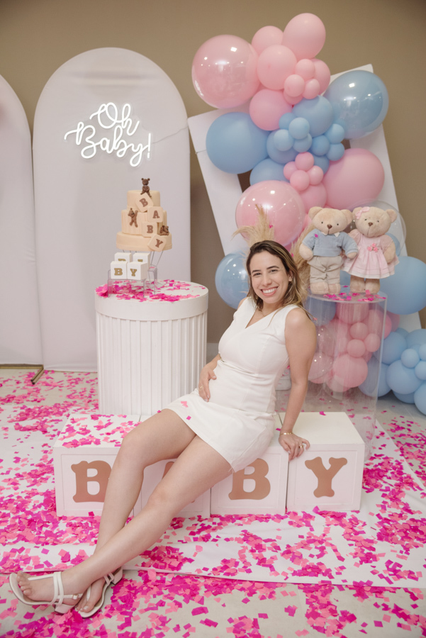 Tamires sorri radiante, sentada sobre cubos decorativos em meio a um tapete de papéis rosa. O cenário de chá revelação, com balões e ursinhos, emoldura a futura mamãe, celebrando com doçura o momento em que a vida da família se tingiu de rosa.