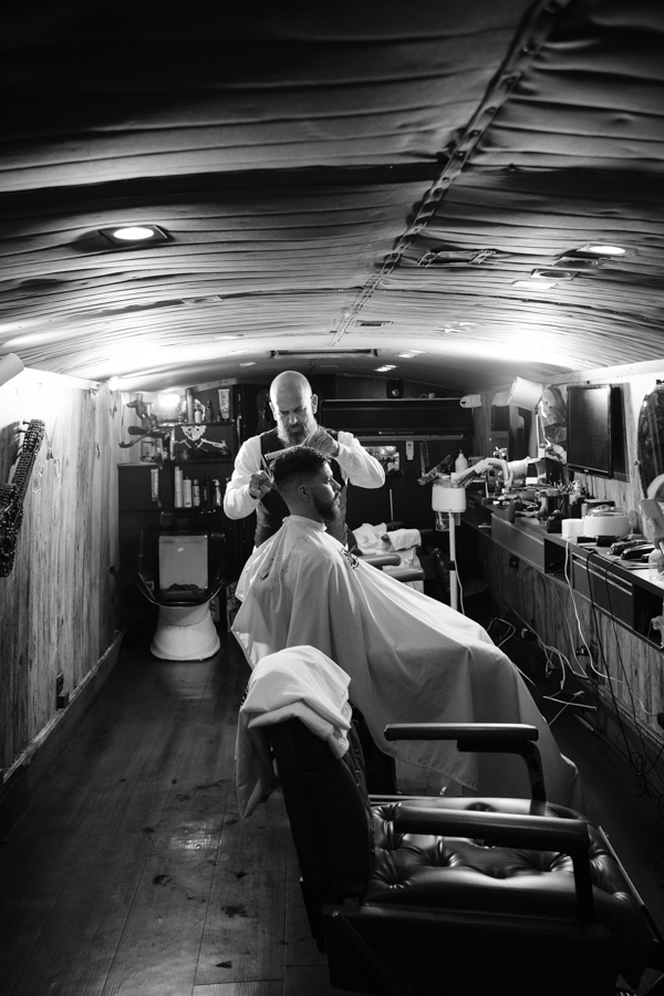 Fotografia de casamento em Jacareí: noivo Matthaus em seu dia de noivo na barbearia. Foto em preto e branco com perspectiva profunda, mostrando o barbeiro finalizando o corte de cabelo em um ambiente rústico e iluminado, capturando a essência do preparati