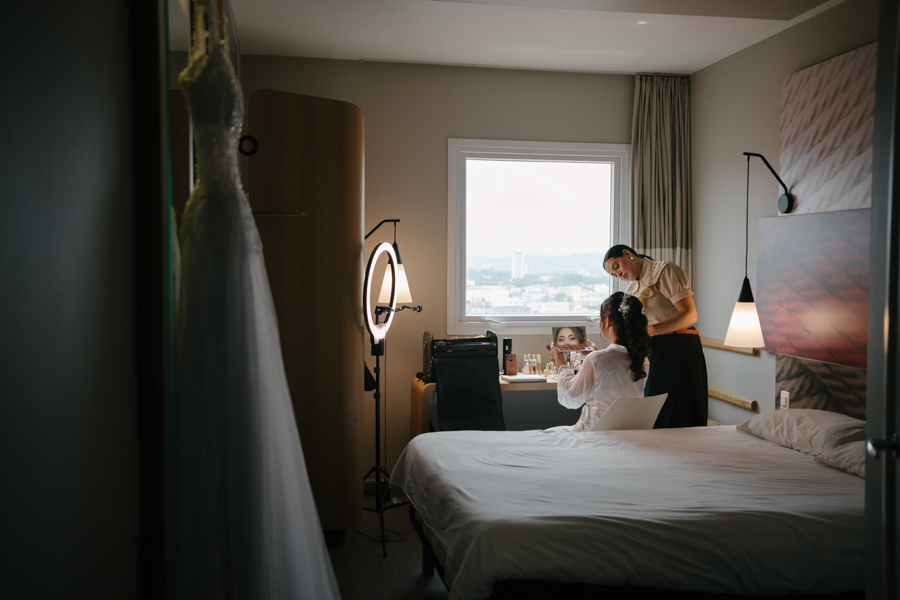Fotografia de casamento em Jacareí: making of da noiva Rebeca no Ibis Hotel. Vista ampla do quarto mostrando o vestido pendurado em primeiro plano, enquanto a noiva finaliza a maquiagem em frente à janela, capturando a luz e o clima da preparação.
