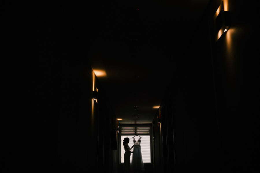 Fotografia de casamento em Jacareí: silhueta artística da noiva Rebeca no corredor do Ibis Hotel. Imagem dramática com luz de fundo vinda da janela, destacando o contorno da noiva e seu vestido pendurado, criando um efeito de luz e sombra único.