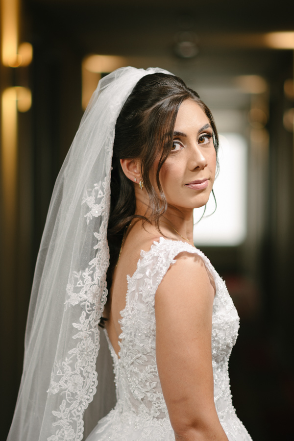 Fotografia de casamento em Jacareí: retrato da noiva Rebeca no Ibis Hotel. Vista de perfil destacando o decote em V nas costas do vestido rendado e o longo véu com bordados delicados, em um registro elegante antes da cerimônia na Chácara Lopes.
