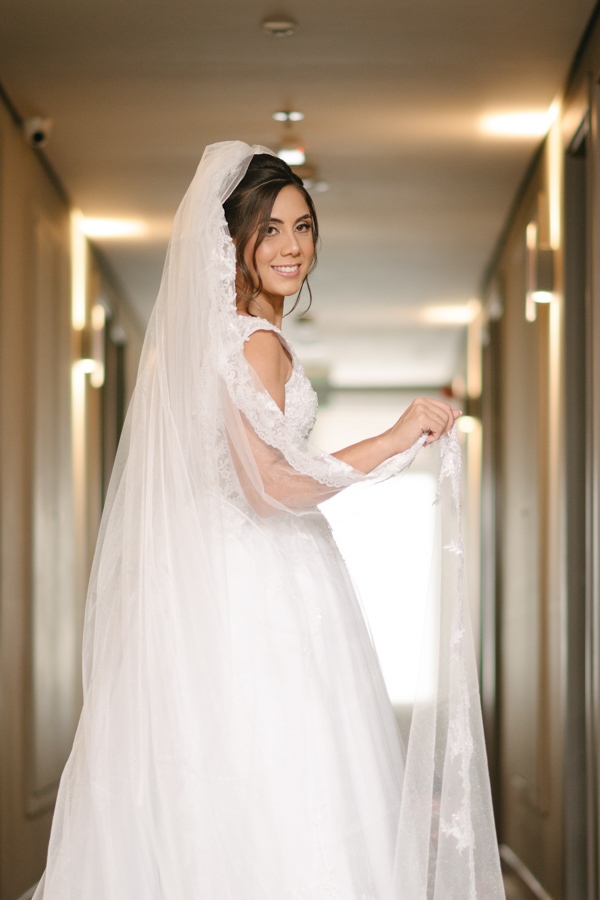 Fotografia de casamento em Jacareí: noiva Rebeca sorrindo no corredor do Ibis Hotel. Foto de corpo inteiro destacando o volume do vestido branco, o longo véu rendado e a felicidade da noiva antes de seguir para a cerimônia na Chácara Lopes.