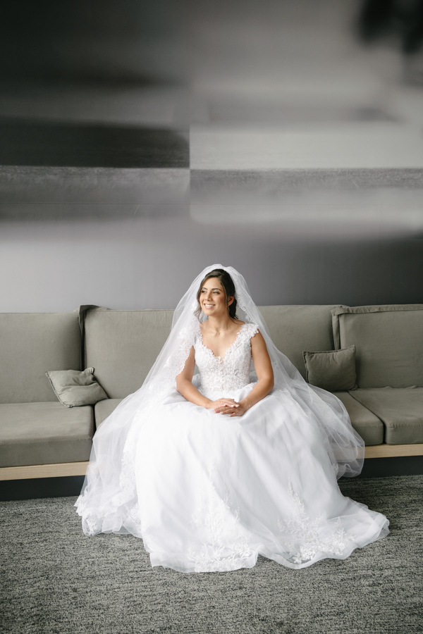 Fotografia de casamento em Jacareí: noiva Rebeca sentada no sofá durante o making of no Ibis Hotel. Registro clássico destacando a plenitude do vestido branco e o longo véu, com a noiva sorrindo serenamente antes da cerimônia na Chácara Lopes.