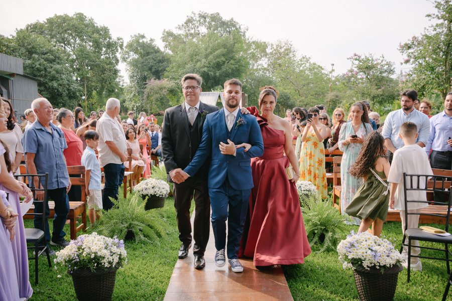 Fotografia de casamento em Jacareí: entrada do noivo Matthaus na Chácara Lopes. O noivo caminha pelo corredor ao ar livre ladeado pelos pais, sob luz natural do entardecer, em uma cerimônia emocionante cercada por convidados e flores brancas.