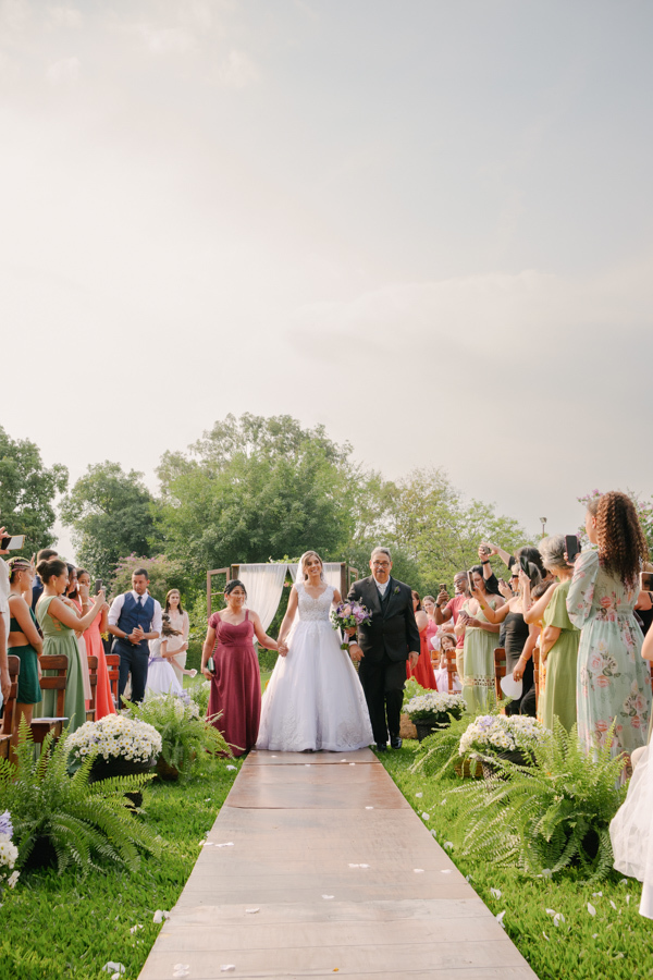 Fotografia de casamento em Jacareí: entrada da noiva Rebeca na Chácara Lopes. A noiva caminha sorridente pelo corredor ao ar livre, acompanhada pelos pais, sob a luz natural do entardecer em uma cerimônia emocionante com convidados e flores.