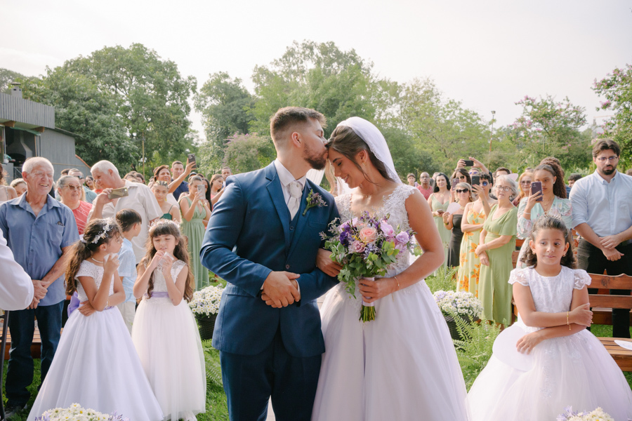 Fotografia de casamento em Jacareí: Matthaus beija a testa de Rebeca no altar da Chácara Lopes. Registro romântico ao ar livre com a noiva segurando o buquê, ladeados por daminhas e convidados sob a luz natural do entardecer.