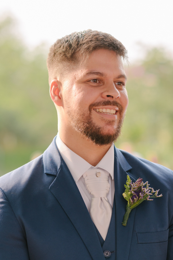 Fotografia de casamento em Jacareí: retrato aproximado do noivo Matthaus no altar da Chácara Lopes. Ele sorri com olhar radiante sob a luz quente do entardecer, vestindo terno azul e lapela de flores roxas durante a cerimônia ao ar livre.