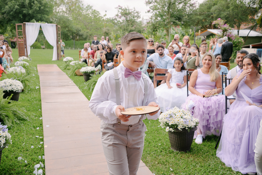 Fotografia de casamento em Jacareí: pajem entrando com as alianças na Chácara Lopes. Menino veste camisa branca, suspensórios e gravata borboleta lilás, caminhando pelo corredor ao ar livre sob o olhar carinhoso dos convidados no entardecer.