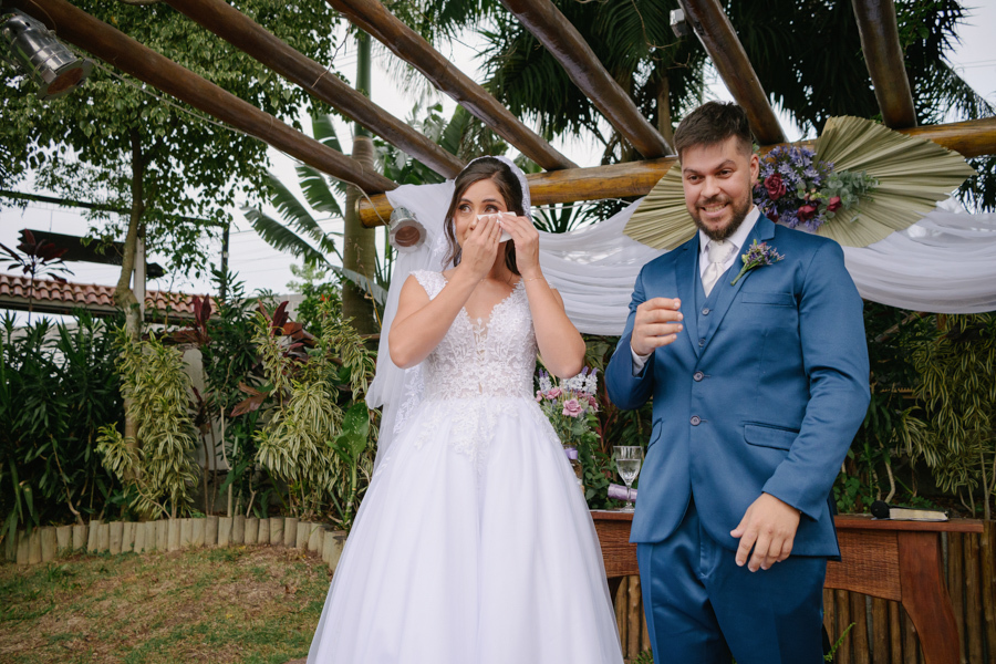 Fotografia de casamento em Jacareí: emoção no altar da Chácara Lopes. A noiva Rebeca enxuga as lágrimas enquanto o noivo Matthaus sorri radiante. Momento espontâneo sob o pergolado rústico decorado, capturando a sensibilidade da cerimônia ao ar livre.